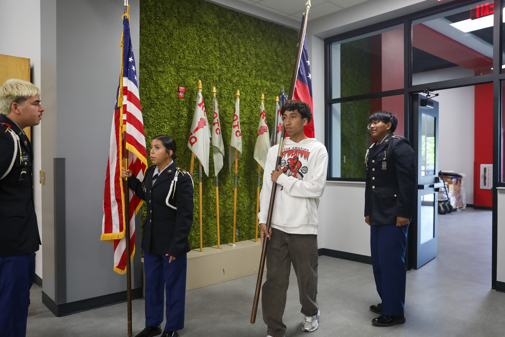 A male JROTC cadet with blonde hair stands facing three other students during color guard practice in a hallway. The students in formation include a female cadet holding the U.S. flag, a male student holding the Georgia flag, and a male cadet holding a ceremonial rifle.