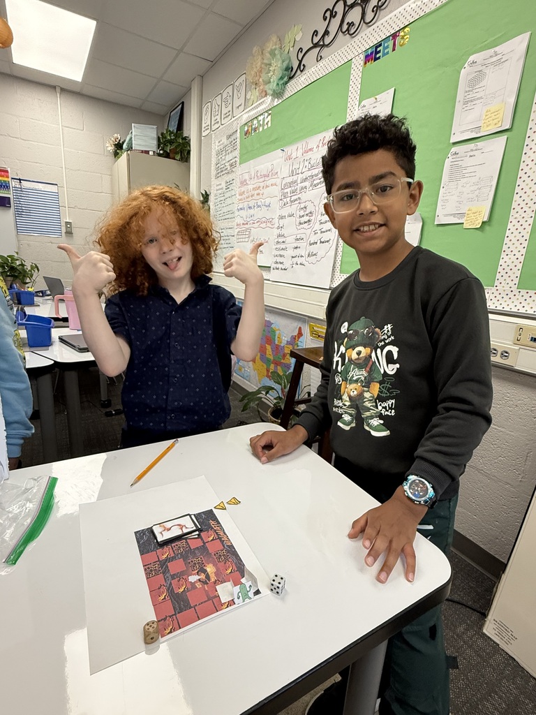 Two male students pose together at a table in a classroom. The student on the left with curly red hair gives two thumbs-up and sticks his tongue out playfully. The student on the right wears glasses and a black sweater while resting his hand on the table. A custom-designed board game printed on paper with red tiles, miniature player tokens, game cards, and a die sits on the white tabletop.