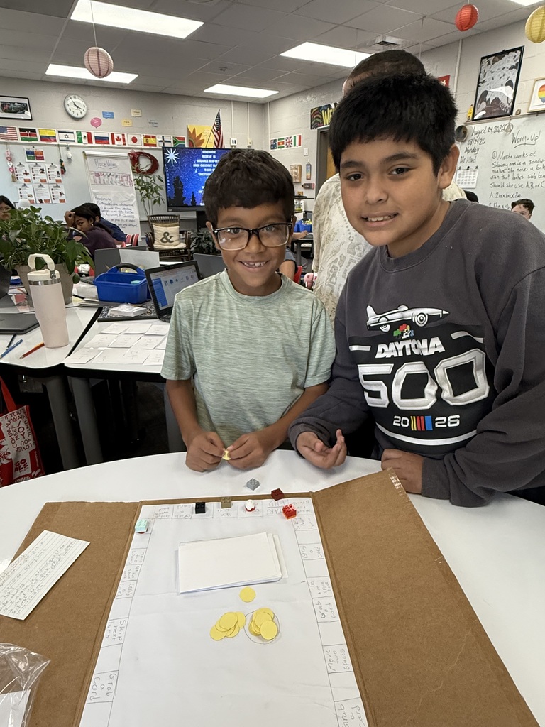 Two young male students stand behind a white classroom table, smiling at the camera. On the table between them is a homemade cardboard board game designed like a square path, featuring Lego bricks as player tokens, paper game cards, and small yellow circular counters stacked in the center.