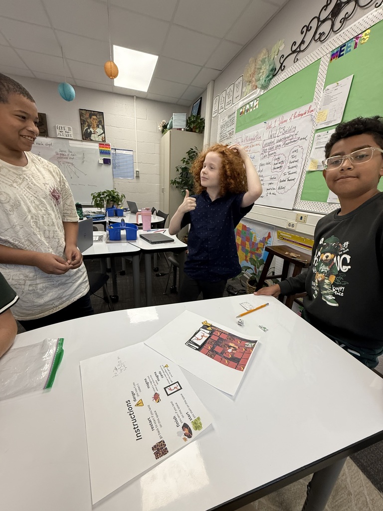 Three students stand around a white table in a classroom during a learning activity. In the center, a student with curly red hair gives a thumbs-up gesture. To the right, a student wearing glasses and a black sweatshirt smiles toward the camera. On the left, another student looks on. On the table rest printed instructions and a custom red-and-black board game design with dice.