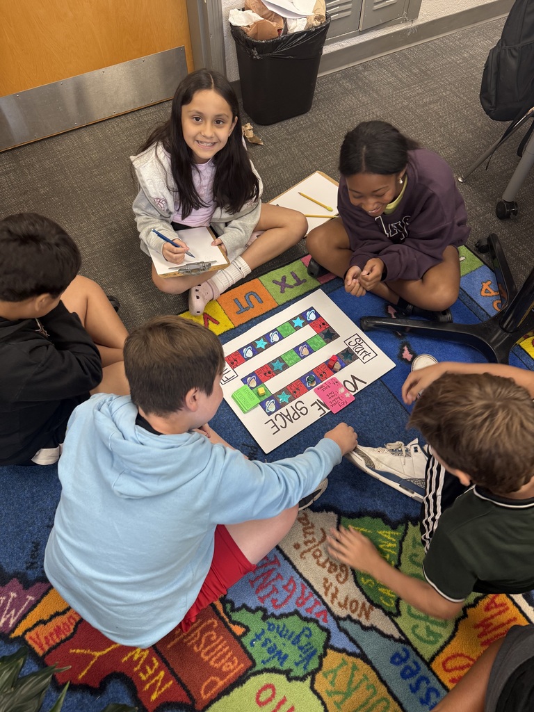 Five elementary students are sitting cross-legged in a circle on a colorful classroom rug patterned with a map of the United States. In the center, they are focused on a handmade tabletop board game titled "SPACE" with colored path tiles, game pieces, and instruction cards. One girl in a grey hoodie holds a clipboard and smiles at the camera, while the other students gather around the board engaging in the activity.