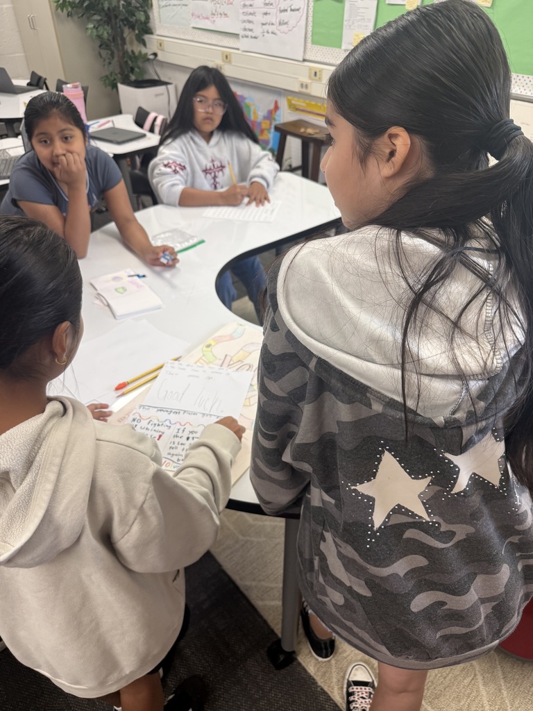 Four female students gather around a curved white table in a classroom setting. Two students in the foreground stand facing the table, looking down at a sheet of handwritten game rules titled "Good Luck" placed beside a colorful drawn game board. Two other students sit across the table, watching and writing on their own papers.
