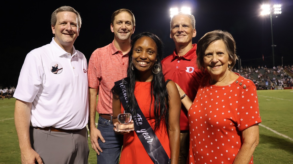 A nighttime photo taken on a illuminated stadium football field featuring five individuals posing together. In the center, a woman wearing a red dress, a black "Teacher of the Year" sash, and dangling earrings holds a clear glass apple award while smiling at the camera. Surrounding her are four colleagues and supporters: a man on the far left in a white polo shirt with a button badge, a man in a red polo shirt, a man in a red Dalton "D" logo shirt, and a woman on the far right in a red polka-dot top.