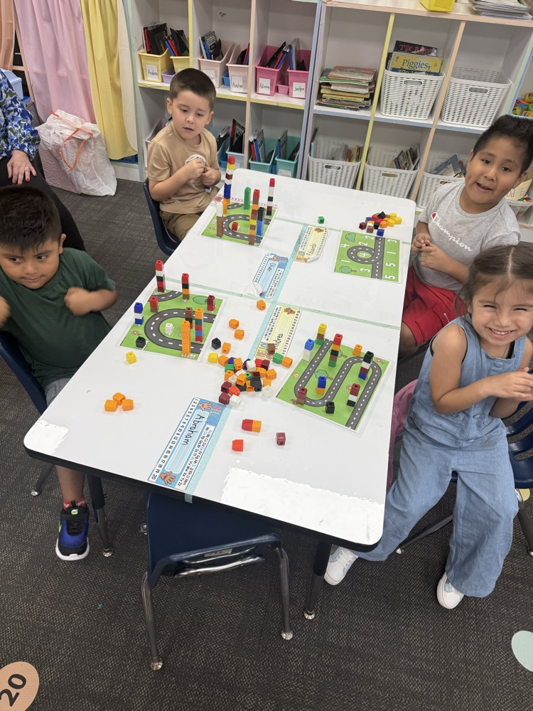 Four young children sit around a white classroom table, smiling as they complete an activity using plastic counting blocks and road-pattern activity mats labeled with student names