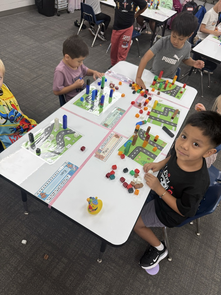 Four elementary students pose happily while gathered around a shared table, engaged in a math or building activity featuring colorful snap-together cubes arranged on laminated activity mats.