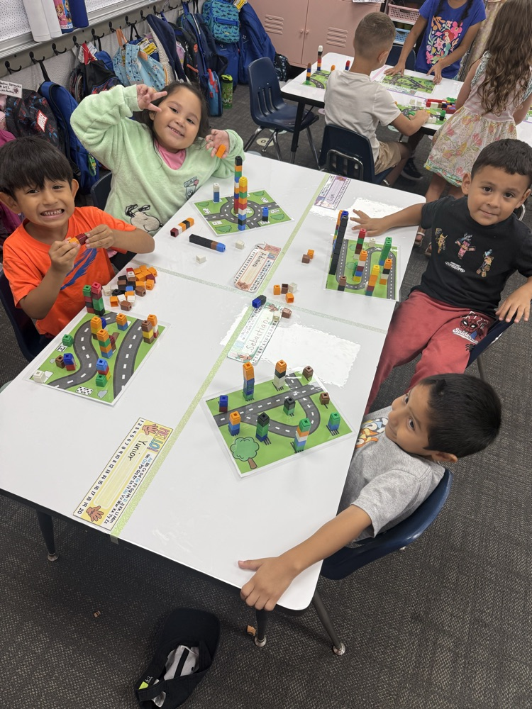 Four children gather around a activity table in a classroom; one girl smiles and flexes her arm playfully while others build patterns using multicolored counting blocks on road-themed work mats.