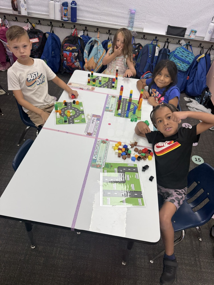 A group of young students sit at a classroom table filled with colorful building blocks, activity mats, and personalized name cards as they work on a learning task.