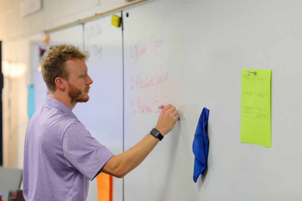 A teacher viewed in profile wears a light purple polo shirt and a smartwatch while writing on a whiteboard with a pink dry-erase marker. A blue cleaning cloth hangs on the board near a green sheet of paper clipped to the surface.