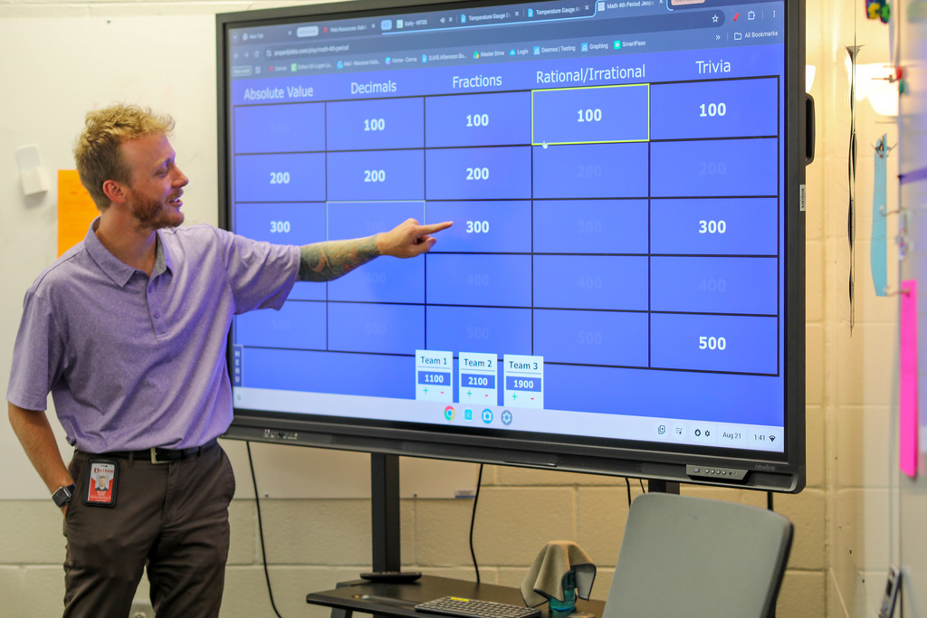 A male teacher wearing a purple polo shirt and ID badge points his finger toward a 300-point square under the "Fractions" category on a digital interactive screen. The screen displays a Jeopardy-formatted math game with categories including Absolute Value, Decimals, Fractions, Rational/Irrational, and Trivia, along with team score counters at the bottom.