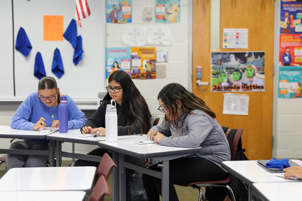 A row of three high school girls sit at gray school desks, leaning over their work and typing into scientific calculators. Educational posters about careers and classroom rules decorate the background wall and door.