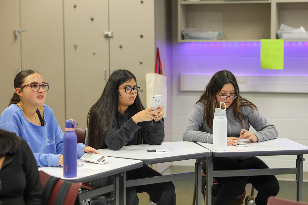 Three high school students sit side-by-side at individual desks in a classroom, each using a handheld scientific calculator. Water bottles sit on their desks, and wall cabinets with ambient under-cabinet lighting are visible in the background.