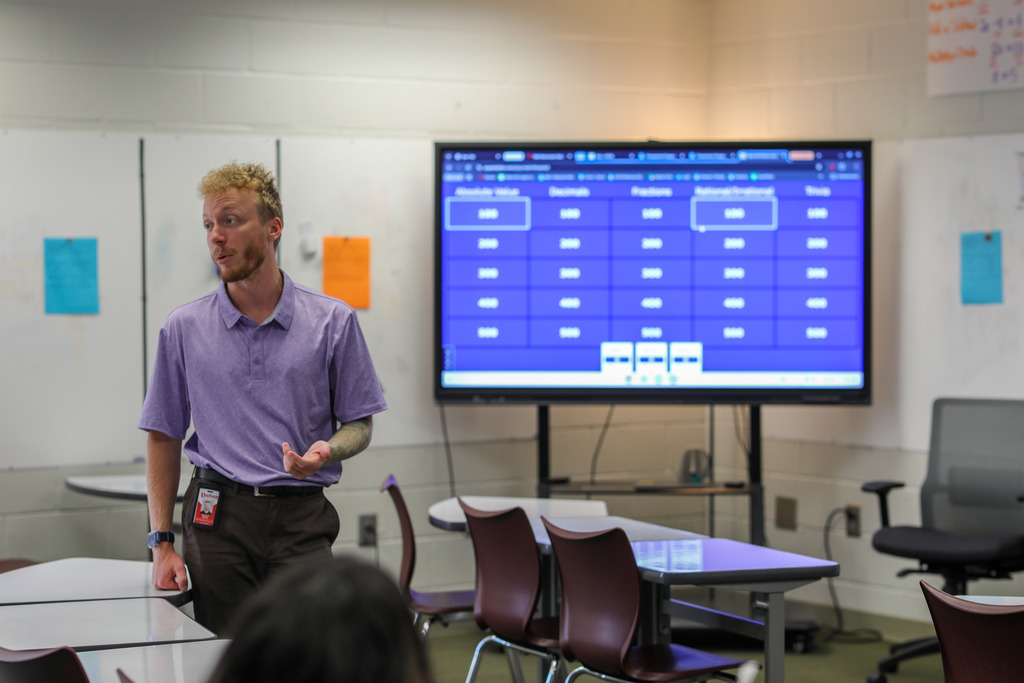 A teacher stands near the front of a room speaking and gesturing open-handed toward students seated at desks. Behind him sits a large display monitor loaded with an educational game board, flanked by whiteboards with colorful paper notes pinned to them.