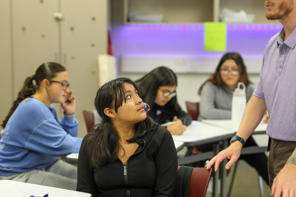A female student wearing a black zip-up jacket sits at her desk, turning her head upward to listen to her teacher standing beside her. Other students in the background focus on their work at neighboring desks.
