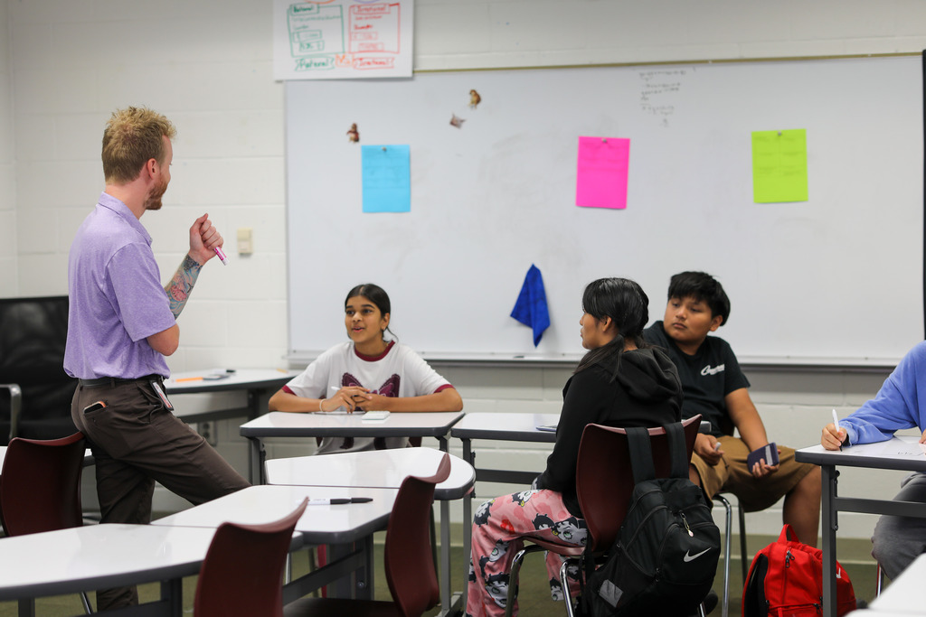 A teacher wearing a purple polo shirt leans against a desk while holding a pink marker and talking with four students seated around him in a semicircle arrangement. A large whiteboard with colorful sticky notes hangs on the wall behind the group.