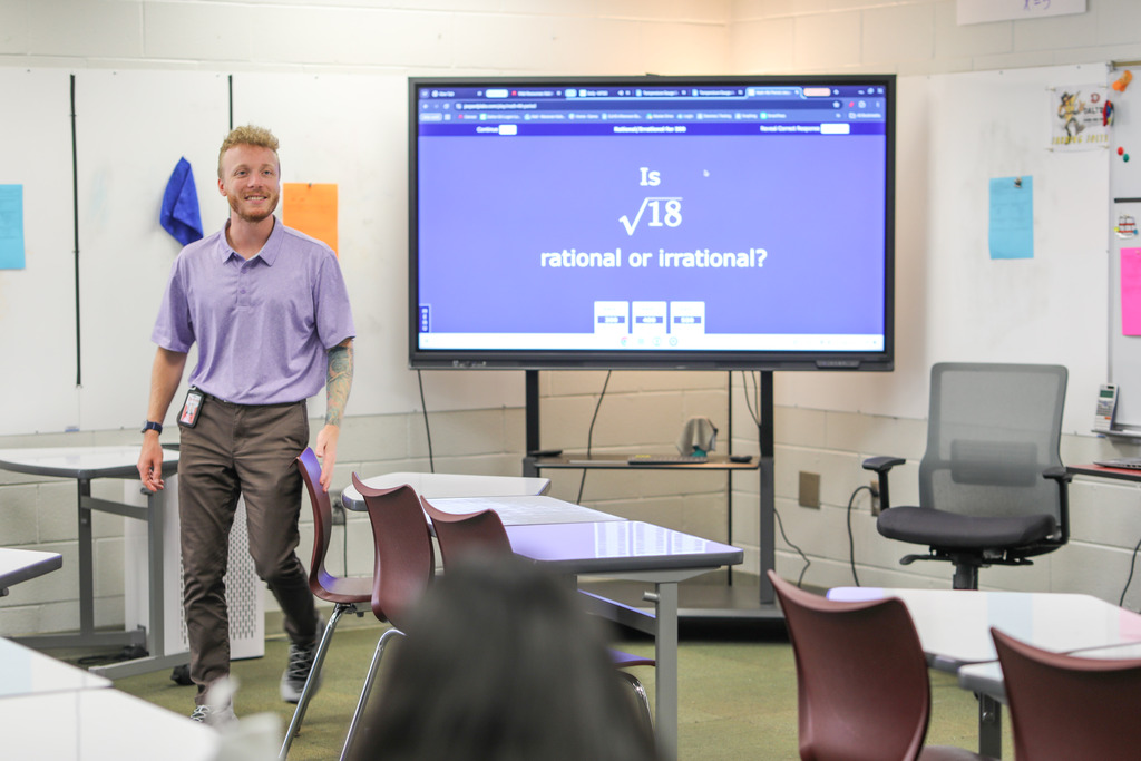 A male teacher in a purple polo shirt and dark trousers smiles while walking between rows of student desks. Behind him, a digital display asks "Is 18 ā rational or irrational?".