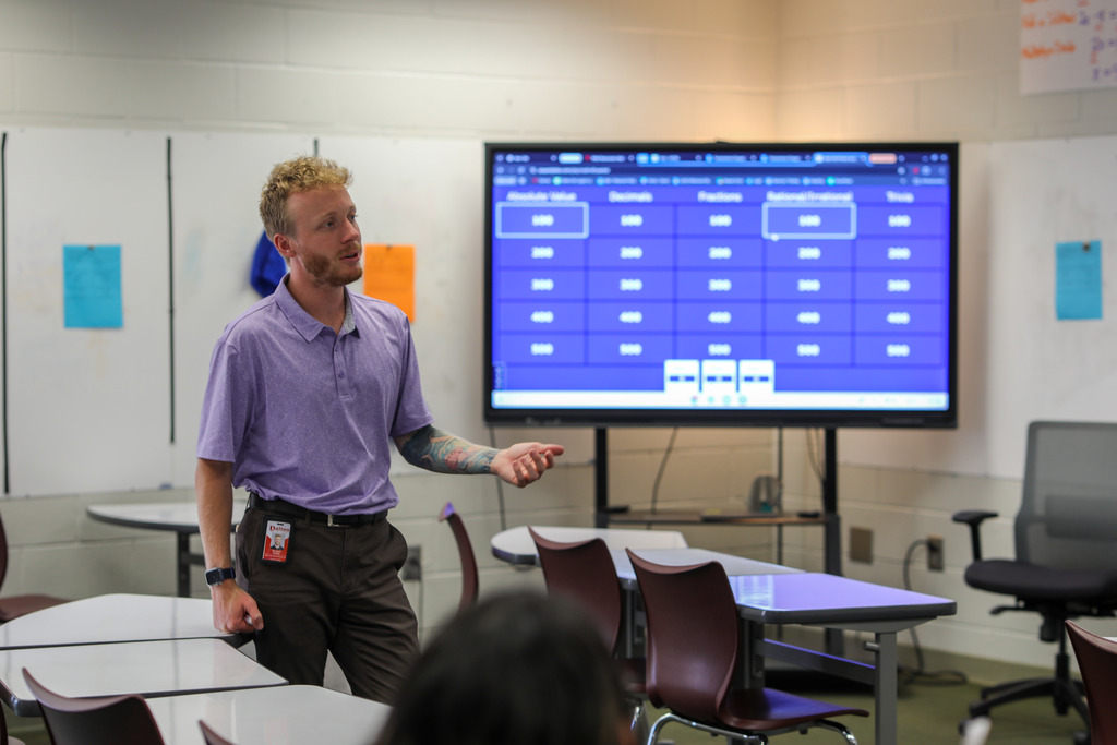 A male teacher with a tattoo on his forearm stands in a classroom speaking to students, holding one hand open in gesture. Behind him, a large flat-panel screen displays a 5x5 blue game board with point values ranging from 100 to 500.