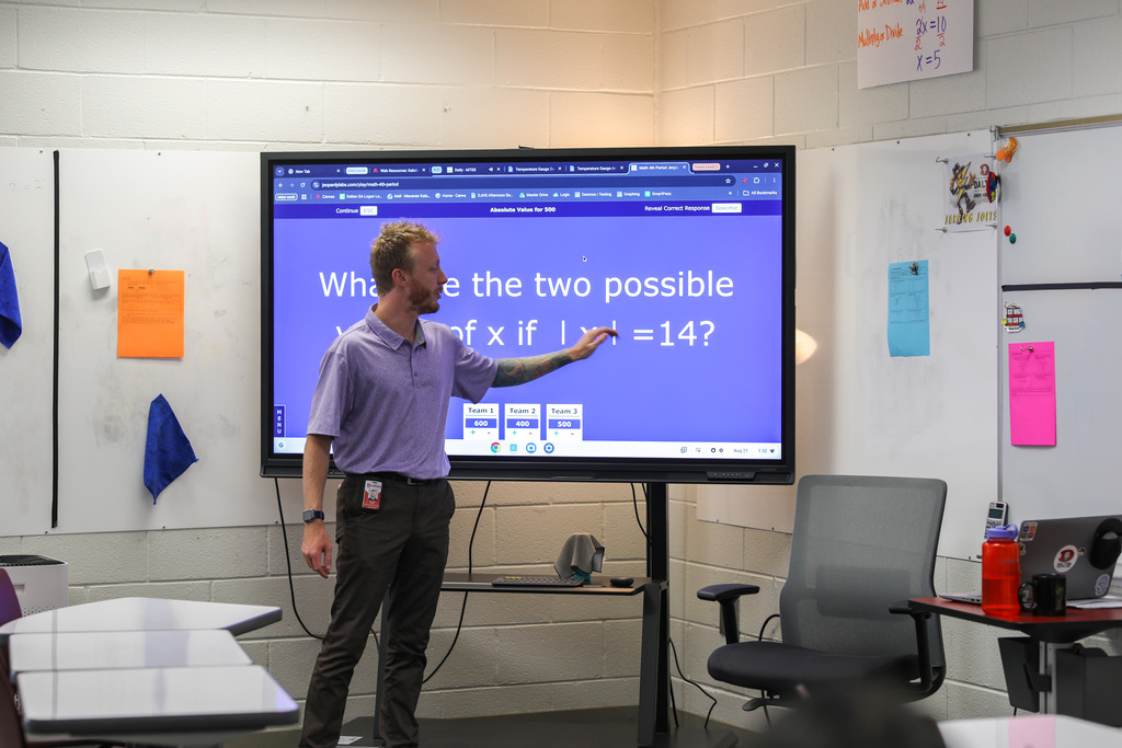 A male teacher stands in front of a large digital display pointing toward a math question reading "What are the two possible values of x if |x| = 14?". Surrounding the display are whiteboards, student desks, an office chair, and classroom supplies.