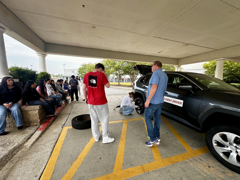 An outdoor Driver's Education demonstration where an instructor in a blue polo stands near a spare tire and instructs a student in a red shirt bending over to use a lug wrench on an SUV's wheel well. A hubcap lies on the ground, and several students sit on a nearby concrete curb watching.