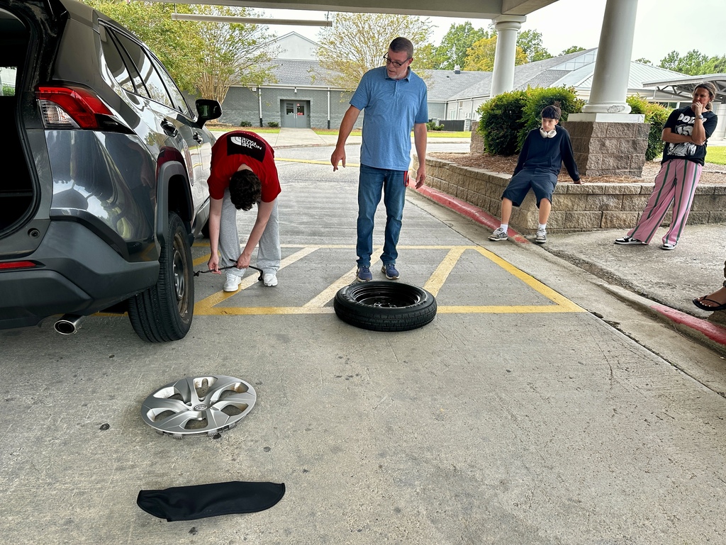 A wide shot of a driver's education practical lesson under a canopy. A female student kneels beside the back wheel of a grey "Student Driver" vehicle to tighten the wheel lug nuts, observed closely by her instructor in a blue polo and a standing classmate in a red shirt, while a row of students watches from a stone ledge.