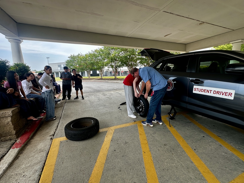 A wide view under a covered walkway where an instructor in a blue shirt and a student work together to align a black spare tire onto a jacked-up dark grey SUV bearing a "STUDENT DRIVER" door sign. A line of high school students sit on a low wall to the left, observing the lesson.