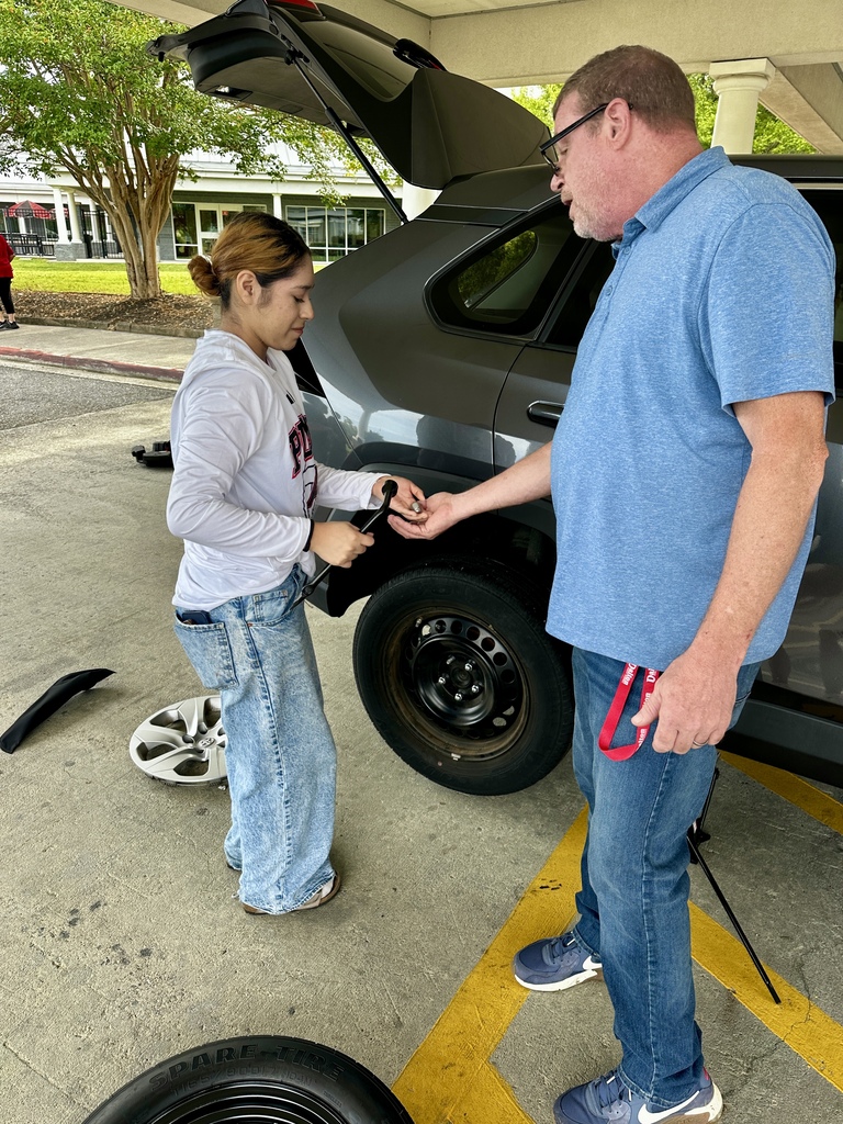 Medium close-up of a female student in a white long-sleeve shirt and light jeans holding a tire iron next to an open trunk and exposed wheel rim. An instructor in a blue shirt hands her lug nuts, with a hubcap and spare tire visible on the ground nearby.