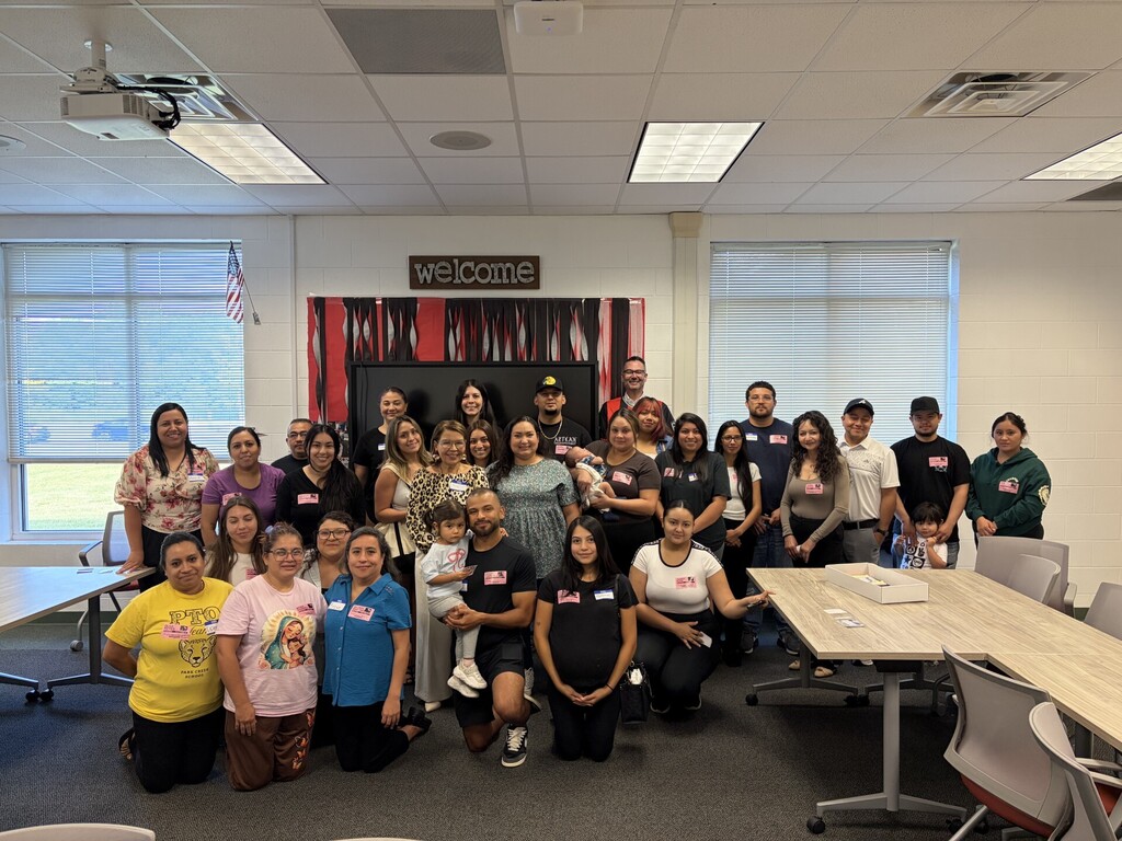 A group of about 25 adults and a young child pose for a group photo inside a classroom setting with large windows. Several participants kneel in the front row while others stand behind them in front of a wall decorated with a "welcome" banner and streamers.