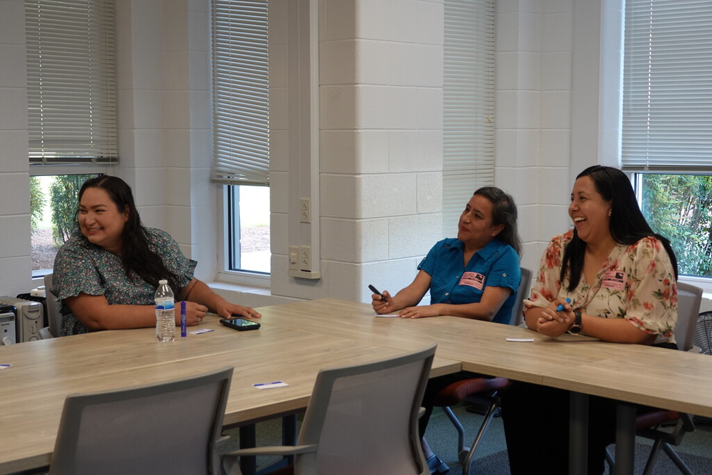 Three women sitting at a table smiling and conversing during a meeting.