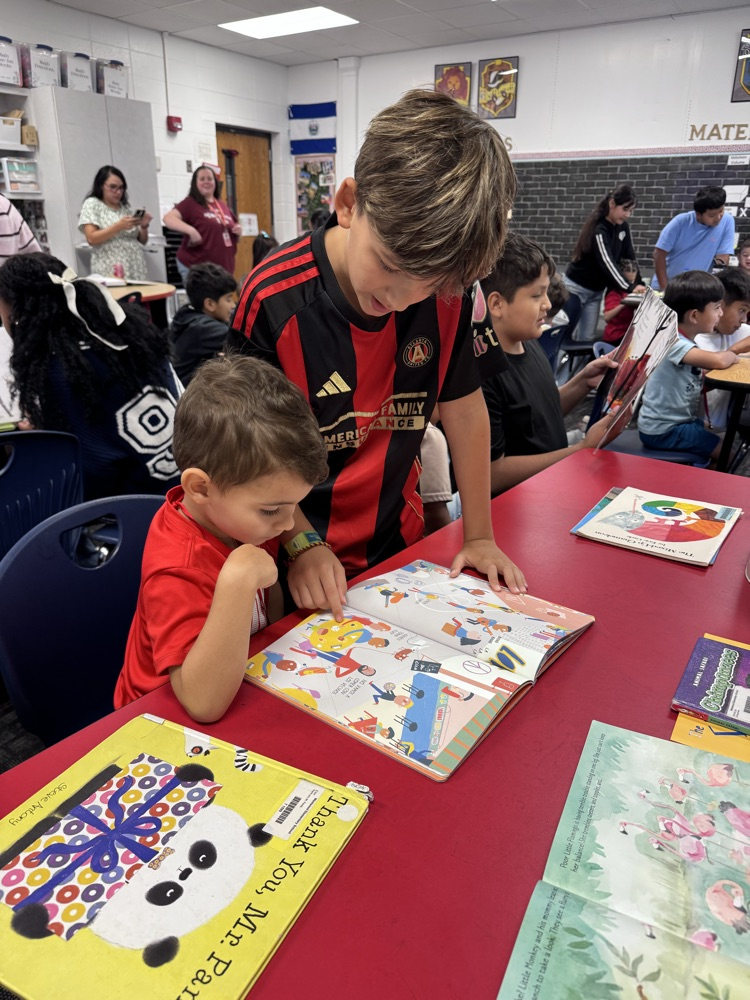 A fifth grade student reads a colorful picture book with a young PreK student at a classroom table while other students read with their buddies nearby