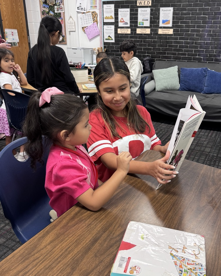 A fifth grade student smiles while reading a book with a young PreK student at a classroom table.