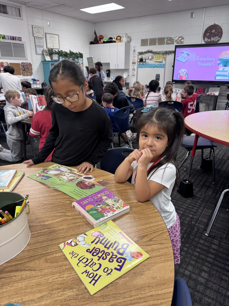 A fifth grade student reads a picture book with a PreK student at a classroom table while other reading buddies work together in the background.