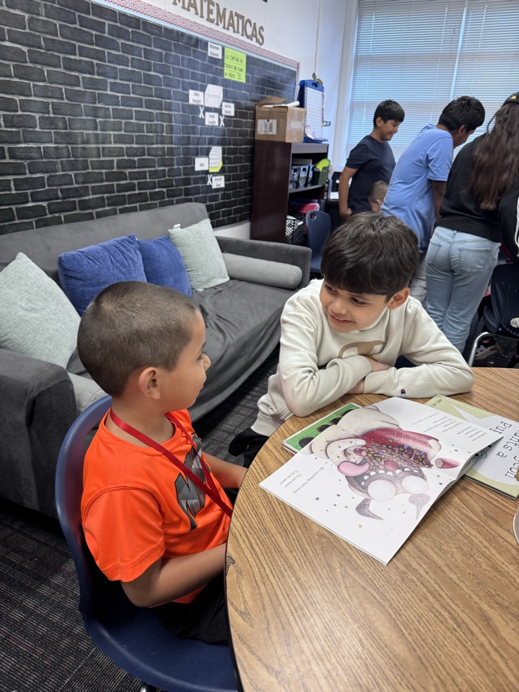 Two students sit across from each other at a classroom table, looking at a picture book together during reading buddy time.