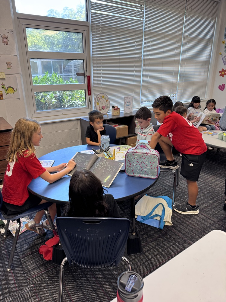 Fifth grade students sit and work with younger reading buddies around a classroom table, sharing books and reading together during Reading Buddies.