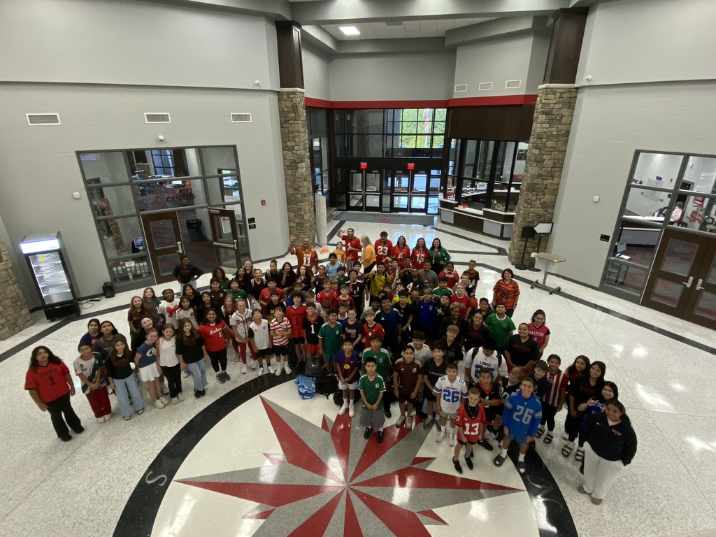 Group photo of sixth grade students participating in HCMS Team Spirit Day. Students are dressed in a variety of team jerseys and sports-themed shirts representing their favorite teams. The group is gathered together in a school hallway, showing off team pride and helping kick off the excitement of football season and fall sports at HCMS.