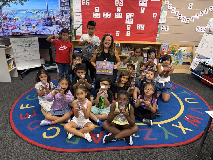 A teacher and group of young elementary students sitting on a colorful alphabet rug in a classroom, posing together while holding small jars with toy goldfish inside. The teacher sits in the center holding the book "We Don't Lose Our Class Goldfish."