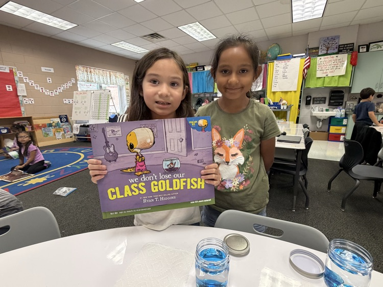 Two young girls standing side by side in a classroom, smiling while holding up the children's picture book "We Don't Lose Our Class Goldfish" by Ryan T. Higgins over a table set with clear jars containing blue liquid.