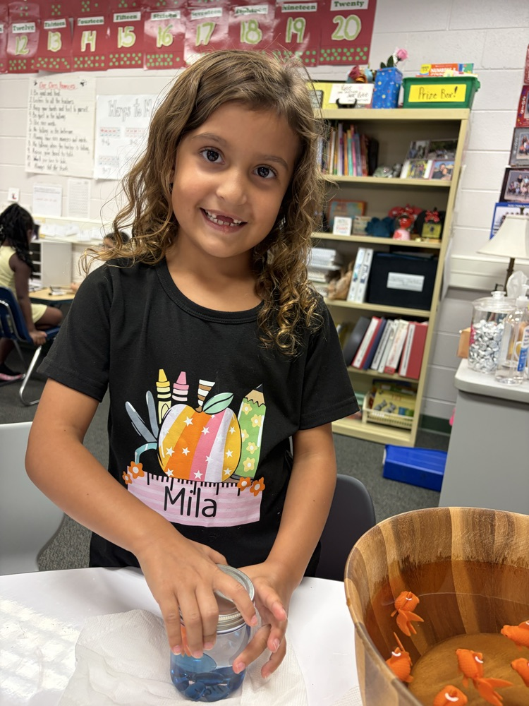 A smiling young girl wearing a black t-shirt putting a metal lid onto a small glass jar containing blue liquid and blue glass pebbles, with a wooden bowl filled with orange toy goldfish on the table nearby.