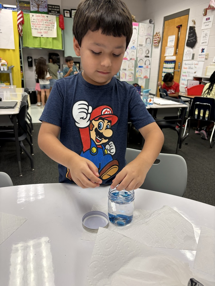 A young boy wearing a blue Super Mario t-shirt standing at a classroom table, focused on placing blue decorative glass gems into a small glass jar filled with water.