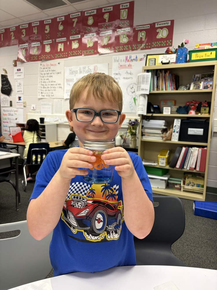 A young boy with glasses and a blue t-shirt with a race car graphic proudly holding up a small glass jar containing blue liquid, blue glass gems, and an orange toy goldfish inside.