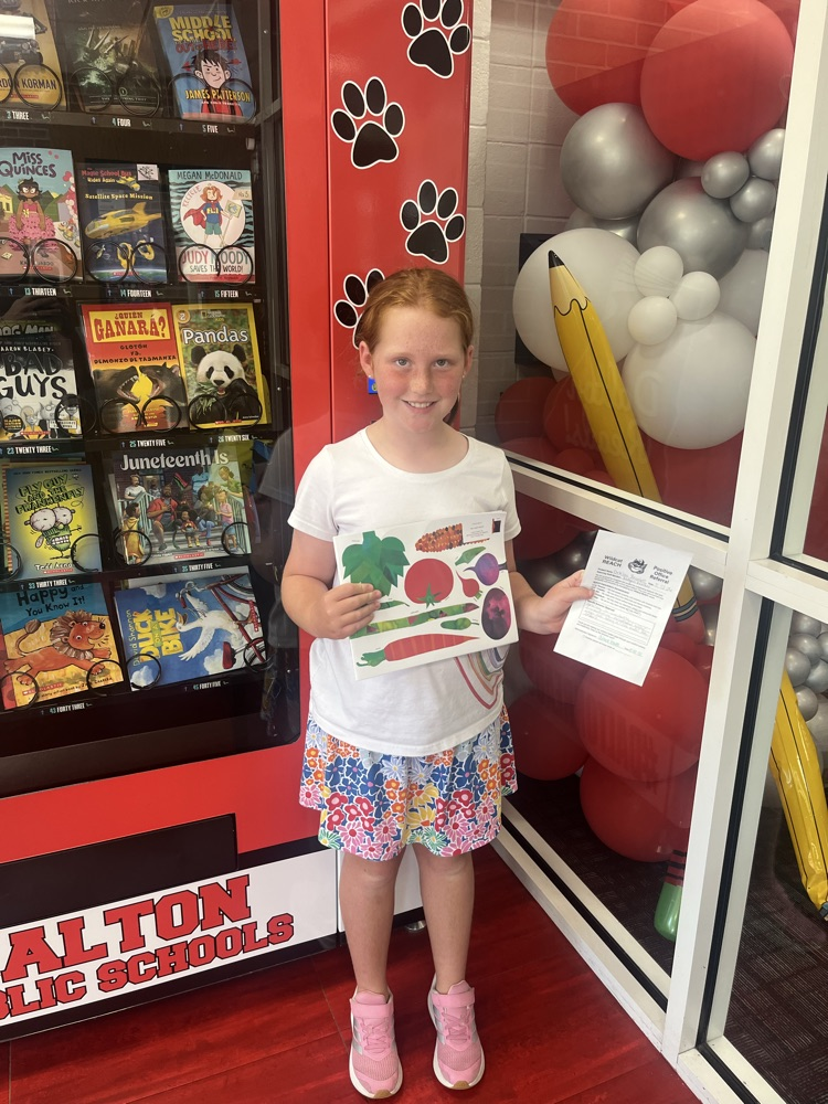 A student smiles while holding a book and Positive Office Referral certificate in front of the Westwood book vending machine.