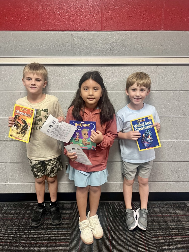 Three students smile while holding books and Positive Office Referral certificates in front of a school wall.