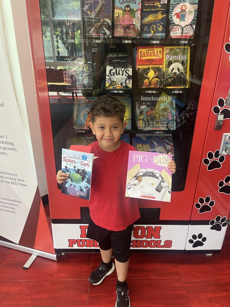 A student smiles while holding two books and a Positive Office Referral certificate in front of the Westwood book vending machine.
