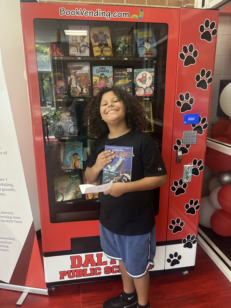 A student smiles while holding a book and Positive Office Referral certificate in front of the Westwood book vending machine.