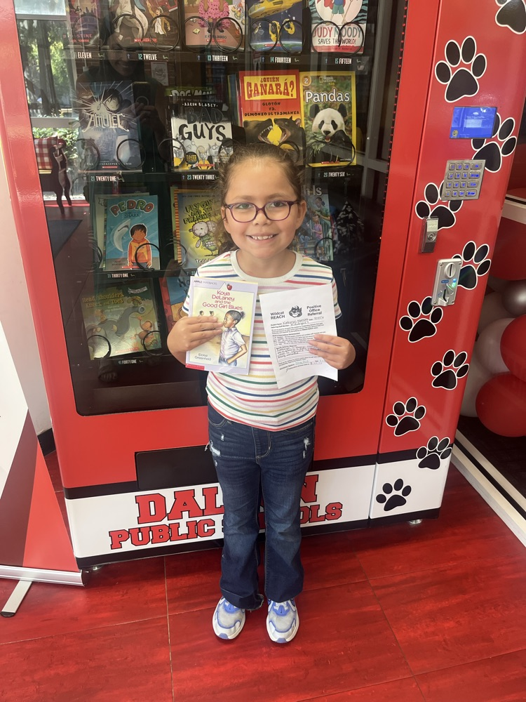 A student smiles while holding a book and Positive Office Referral certificate in front of the Westwood book vending machine.