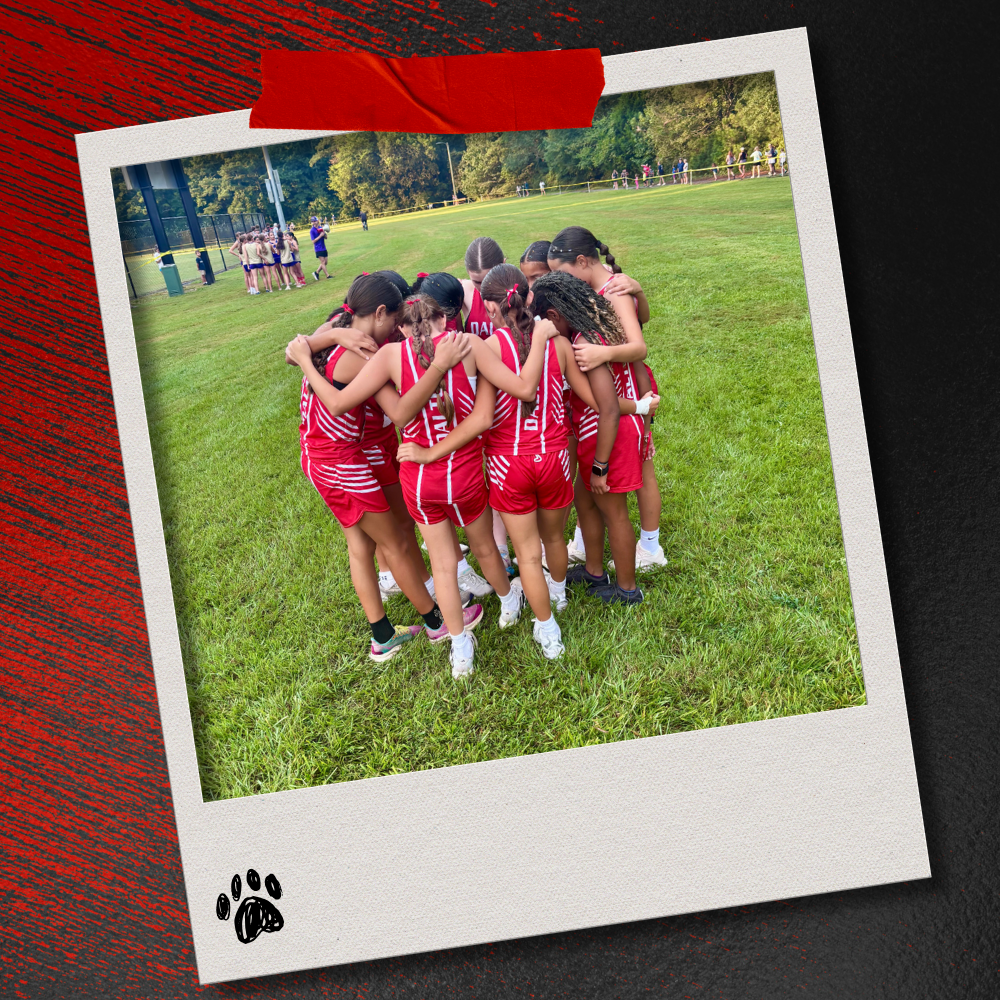 Dalton Middle girls cross country runners huddle together with their arms around one another before the race on a grassy course. The photo is displayed in a taped Polaroid-style frame on a textured red-and-black background with a paw print accent.