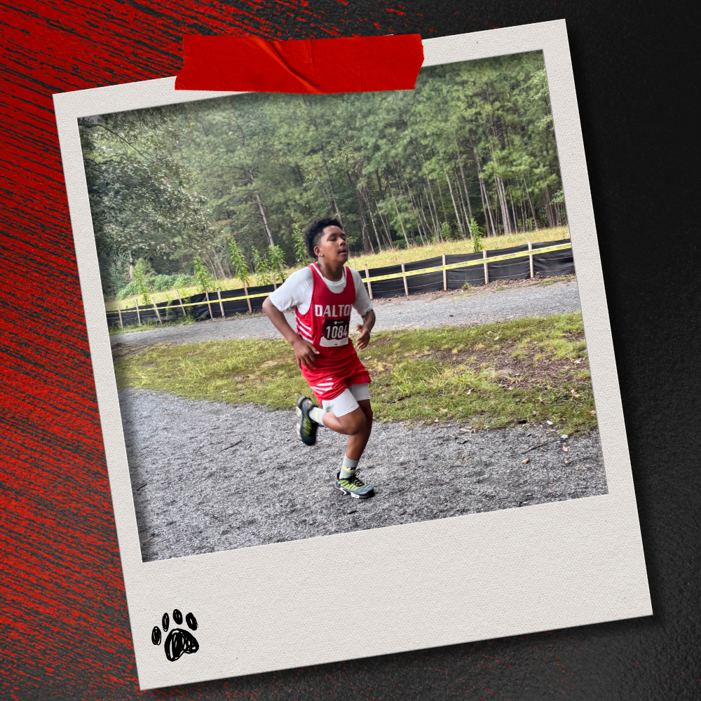 A Dalton Middle boys cross country runner in a red uniform competes on a gravel section of the course surrounded by trees. The photo is displayed in a taped Polaroid-style frame on a textured red-and-black background with a paw print accent.