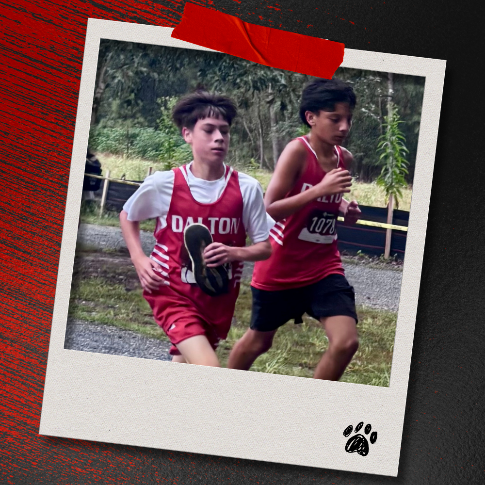 Two Dalton Middle boys cross country runners in red uniforms run alongside one another during the race. The photo is displayed in a taped Polaroid-style frame on a textured red-and-black background with a paw print accent.