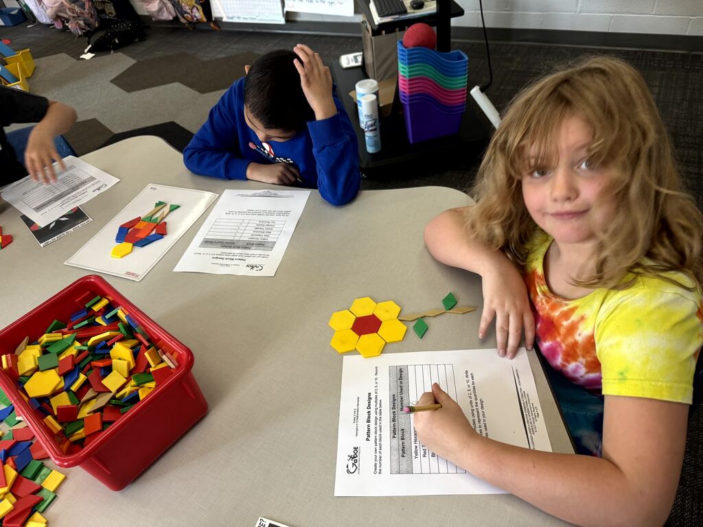 A blonde girl wearing a tie-dye shirt smiles at the camera while sitting at a classroom table. On her worksheet, she has arranged yellow hexagons, a red trapezoid, and green triangles to create a flower design. In front of her sits a red bin full of pattern blocks, and another student works in the background.