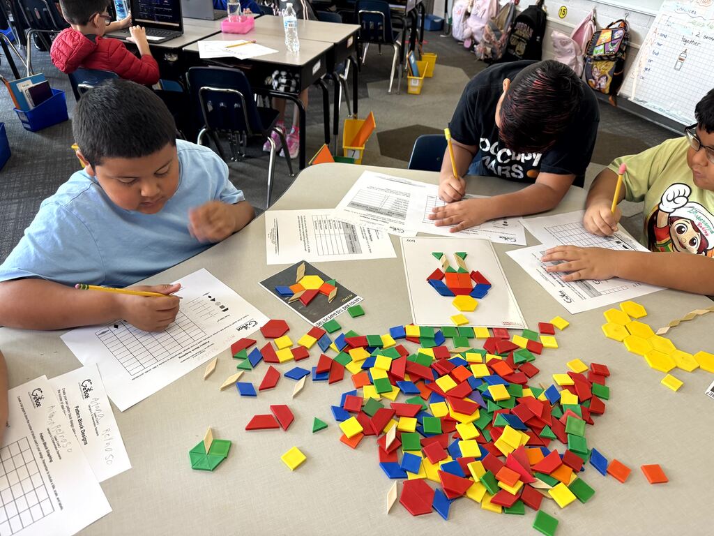 Students sit around a table working on a pattern block assignment. A girl in a pink shirt writes on her worksheet on the left, while a boy in a light blue shirt rests his head on his hand on the right. A template worksheet showing a bunny shape made of blocks rests on the table, alongside scattered plastic pattern shapes.
