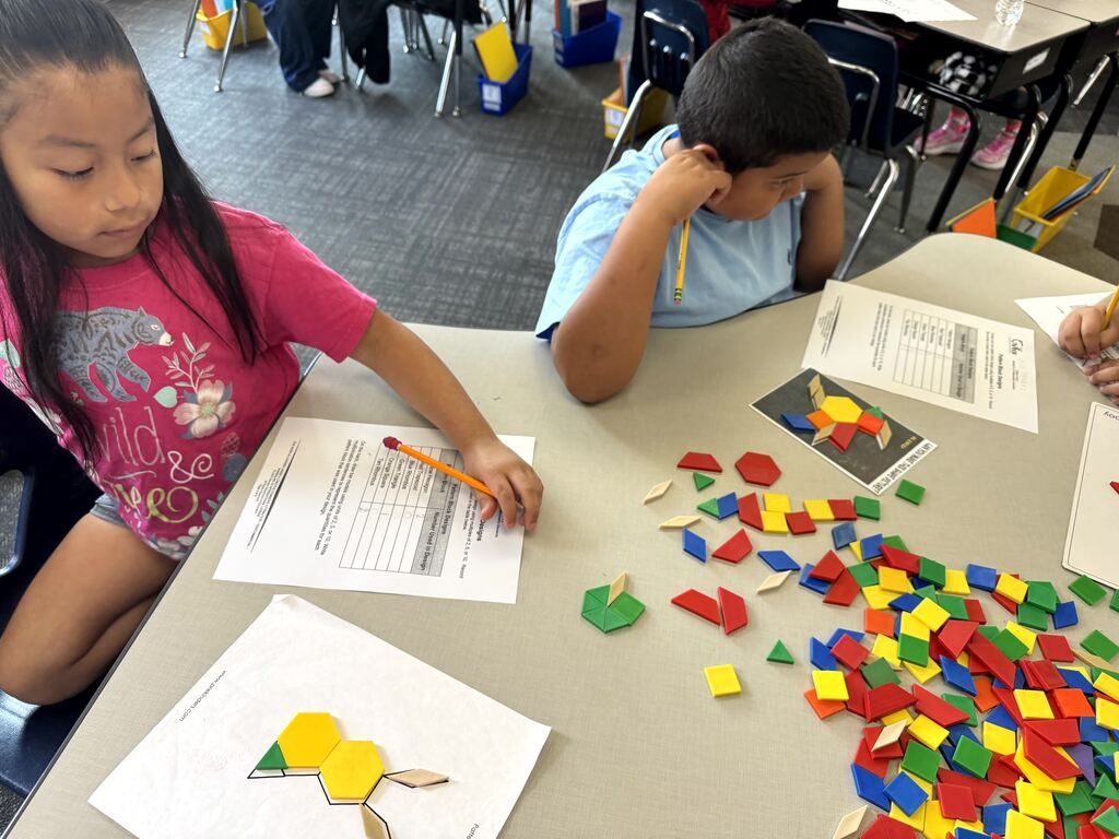 A young girl with dark hair wearing a pink shirt and hair bow sits at a classroom table, holding a pencil and filling out a graph worksheet. Beside her paper is a template showing a shape design created with yellow, green, and tan pattern blocks, along with a large pile of colorful plastic blocks on the table.