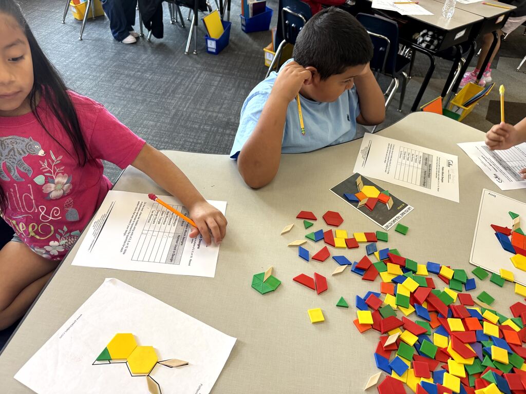 Students sit around a table working on a pattern block assignment. A girl in a pink shirt writes on her worksheet on the left, while a boy in a light blue shirt rests his head on his hand on the right. A template worksheet showing a bunny shape made of blocks rests on the table, alongside scattered plastic pattern shapes. Image 6 (IMG_4956.jpg)